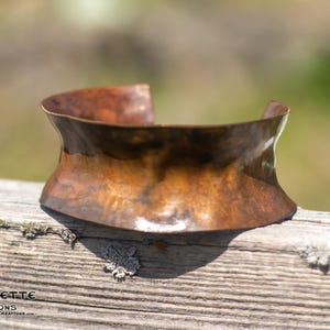 May include: A wide copper bracelet with a hammered finish. The bracelet is curved and has a slightly irregular shape.