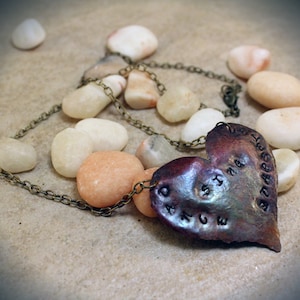 Puede incluir: Un collar con colgante en forma de corazón con las palabras "ANGE HOPE" y "ST. JAMES" grabadas en la parte delantera. El colgante es una mezcla de tonos bronce y morado, y está rodeado de pequeñas piedras lisas. La cadena del collar es de color bronce.