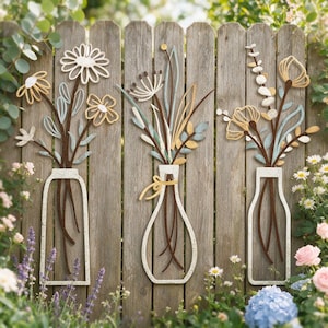 May include: Three wall-mounted metal art pieces featuring stylised floral arrangements in vases. Each piece displays thin, wire-like stems and petals in shades of white, light blue, and mustard yellow, creating a cohesive set of botanical wall decor.