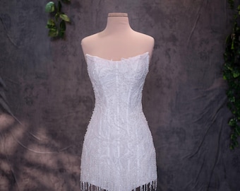 May include: A strapless white mini dress featuring an intricate all-over pattern of beads and sequins. The garment has a fitted bodice and a hemline finished with a delicate fringe of hanging beads that create a textured, shimmering appearance.