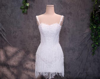 May include: A white mini dress featuring a sweetheart neckline, thin straps, and an all-over lace overlay. The bodice is embellished with intricate beadwork, and the skirt is finished with a long, layered fringe hem that hangs from the waist.