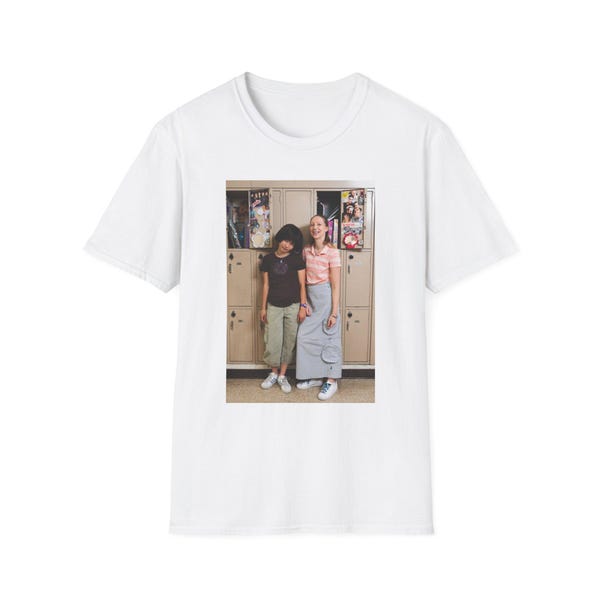 Op de afbeelding: Een wit t-shirt met korte mouwen en een gecentreerde rechthoekige grafische print. De afbeelding toont twee personen die voor beige metalen kluisjes staan en vrijetijdskleding dragen, waaronder een donkere top, een gestreept shirt, een broek en een rok.