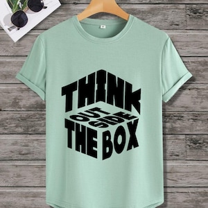 May include: A light green short-sleeved t-shirt with a crew neckline. The center of the shirt features a black graphic design of a cube with the text "THINK OUTSIDE THE BOX" printed in a bold, sans-serif font that follows the cube's perspective.