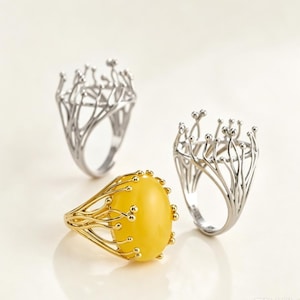 Op de afbeelding: Drie ringen met een complexe, takachtige metalen zetting met kleine bolvormige uiteinden. Twee ringen zijn van zilverkleurig metaal, terwijl de derde ring goudkleurig is en een grote, gladde, ovaalvormige gele steen in het midden heeft.