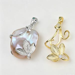 May include: A silver-toned pendant featuring an irregular iridescent pearl accented with a textured metal flower and a small clear stone, alongside a gold-toned metal pendant shaped like a swirling vine with a textured flower and clear stone.