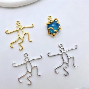 May include: A collection of jewellery charms including two silver-toned and one gold-toned wire-frame stick figure pendants, alongside a gold-toned charm featuring a round, translucent blue bead encased in a decorative, swirling metal cage design.