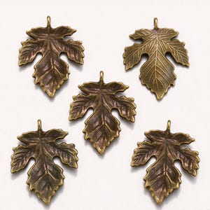 Puede incluir: Cinco dijes de metal en tono bronce antiguo con forma de hoja, dispuestos en grupo. Cada dije presenta un patrón de venas en relieve detallado y un pequeño lazo circular en la parte superior para su fijación. El metal tiene un acabado oscuro.