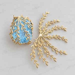 Puede incluir: Un conjunto de accesorios para el cabello en metal dorado que incluye una gran piedra ovalada de color azul claro facetada dentro de una delicada jaula de hojas doradas, junto a una peineta de rama dorada con pequeños cristales redondos.