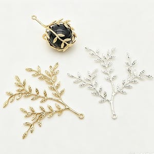 May include: A gold-toned metal cage pendant containing a smooth black spherical bead, accompanied by two flat metal branch-shaped charms, one in a gold finish and one in a silver finish, each featuring detailed leaf patterns and small loops.
