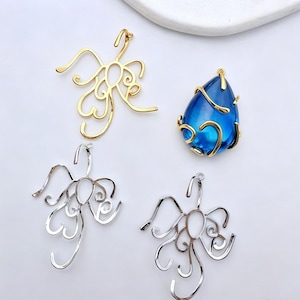 Puede incluir: Tres colgantes de alambre metálico y un colgante de cristal azul en forma de lágrima envuelto en alambre dorado. Dos de los colgantes son plateados y uno es dorado. Cada pieza de alambre presenta un diseño abierto, ondulado y abstracto.