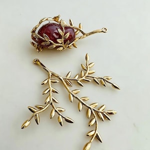 Puede incluir: Dos componentes de joyería de metal en tono dorado. Una pieza es un colgante con forma de rama con hojas pequeñas. La segunda pieza es una estructura de rama dorada similar que envuelve una piedra ovalada lisa de color rojo intenso.