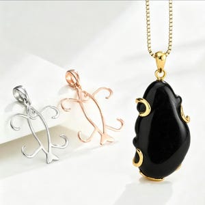 May include: A gold-toned necklace featuring a smooth, polished black stone pendant with gold accents, accompanied by two separate metal fish-shaped pendants in silver and rose gold tones, each with a bail for attachment to a chain.