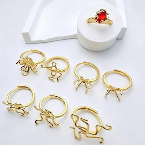 May include: A collection of gold-toned metal rings featuring various abstract, wire-like designs. One ring is set with a prominent oval red gemstone, while the others are empty settings with intricate, curved metal prongs and open bands.
