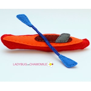 May include: A felt toy kayak with a blue paddle. The kayak is red and orange with a gray seat. The toy is handmade and has a stitched design.