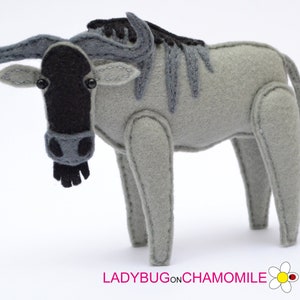 May include: A gray felt stuffed animal wildebeest with black horns, eyes, and tail. The animal is standing on a white surface.