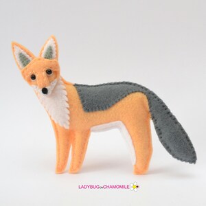 May include: A felt fox figurine featuring orange, white, and gray sections with visible stitched edges. The fox has black bead eyes and a black nose. The text LADYBUGONCHAMOMILE with a small flower icon is printed at the bottom of the image.