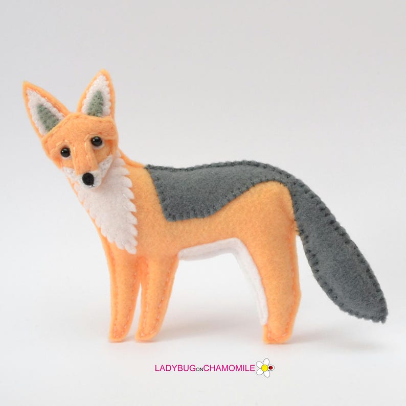 May include: A felt fox figurine featuring orange, white, and gray sections with visible stitched edges. The fox has black bead eyes and a black nose. The text LADYBUGONCHAMOMILE with a small flower icon is printed at the bottom of the image.