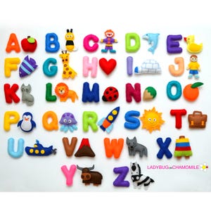 Op de afbeelding: Een set kleurrijke vilten alfabetletters met bijbehorende objecten, waaronder een appel, bij, clown, dolfijn, eend, vis, giraf, hart, iglo, persoon, kat, leeuw, lieveheersbeestje, noot, sinaasappel, pinguïn, octopus, raket, zon, aktetas, onderzeeboot, vulkaan, wolf, speelgoed, jak en zebra.
