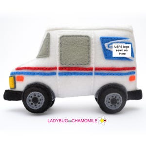 May include: A white felt mail truck ornament featuring a blue and red horizontal stripe, grey windows, black wheels with grey centers, and a blue square patch with the text USPS logo sewn on Here. The truck has a yellow headlight and orange marker.