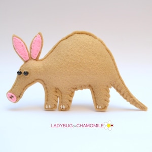 Può includere: Un giocattolo di formichiere in feltro marrone con orecchie, naso e dettagli rosa. Il formichiere ha occhi di perline nere e dettagli bianchi sulle zampe. Il giocattolo è fatto a mano e ha un design stravagante. Il testo "LADYBUGonCHAMOMILE" è in basso.