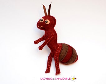 May include: A handcrafted felt ant figurine in deep red with brown abdominal stripes, featuring stitched details, two brown ribbon antennae, and two small black bead eyes. The text LADYBUGONCHAMOMILE with a small flower icon is printed below.