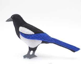 May include: A felt magpie bird ornament featuring a black head, back, and tail, with a white chest and wing patch, and a vibrant blue wing section. The bird has grey feet and a grey beak, with the text LADYBUGONCHAMOMILE printed below the figure.