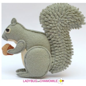 Könnte beinhalten: Eine Eichhörnchen-Figur aus Filz in Salbeigrün und Creme, die eine kleine braune Eichel hält. Das Eichhörnchen hat einen strukturierten, zotteligen Schwanz aus einzelnen Filzschlaufen und sichtbare Nähte. Darunter steht LADYBUG ON CHAMOMILE.