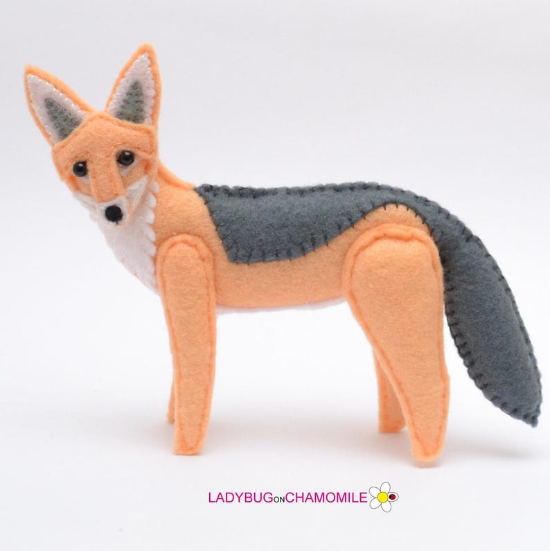 May include: A felt fox figurine featuring a light orange body with a white chest and face, dark gray accents on the ears, back, and tail, and black bead eyes. The text LADYBUGONCHAMOMILE with a small flower icon is printed at the bottom.
