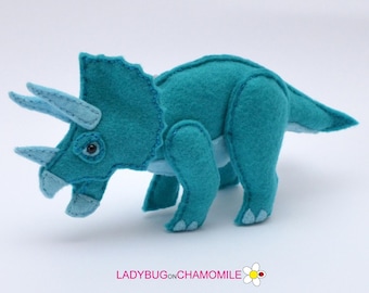 May include: A teal felt triceratops dinosaur toy with light blue horns and accents. The dinosaur features visible stitched seams, a large frill, and a small dark eye. Below the toy, the text LADYBUGONCHAMOMILE is printed with a flower icon.