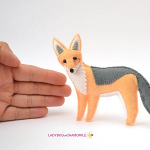 May include: A felt fox figurine featuring a light orange body, a white chest and face, and dark gray accents on the back, tail, and ears. The fox has small black eyes and a black nose. The text LADYBUGONCHAMOMILE is printed at the bottom.