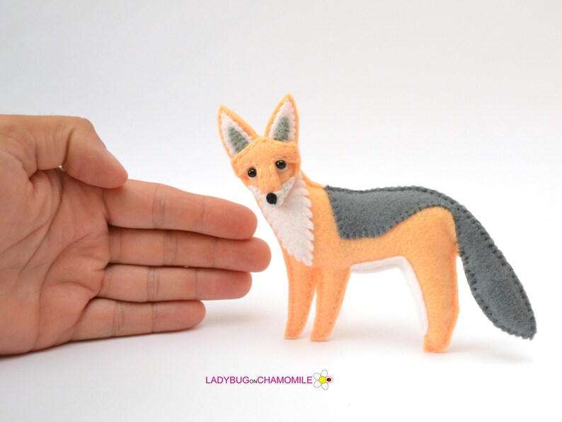 May include: A felt fox figurine featuring a light orange body, a white chest and face, and dark gray accents on the back, tail, and ears. The fox has small black eyes and a black nose. The text LADYBUGONCHAMOMILE is printed at the bottom.