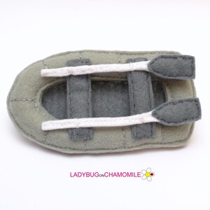 May include: A gray felt toy boat with white felt ropes and two gray felt oars. The boat is designed to look like a small inflatable raft.