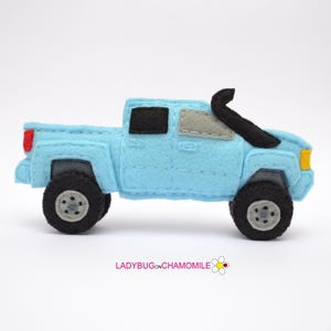 May include: A light blue felt toy truck with black wheels and a black snorkel. The truck has a black window and a gray window. The truck has a red tail light and a yellow headlight. The toy truck is a handmade item.