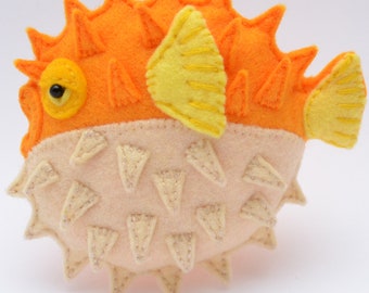 Puede incluir: Un juguete de pez globo de fieltro hecho a mano, con fieltro naranja y amarillo. El pez globo tiene una barriga rosa con púas de fieltro blanco.