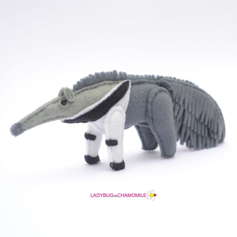 Puede incluir: Un juguete de oso hormiguero de fieltro hecho a mano en tonos grises, blancos y negros. El oso hormiguero tiene un hocico largo, un cuerpo gris y un pecho y patas blancos con detalles negros. El juguete est&aacute; sobre una superficie blanca.