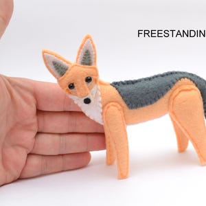May include: A handcrafted felt fox figurine featuring light orange fur, a white chest, and dark gray accents on the back, ears, and tail. The fox has black bead eyes and a black nose, with visible stitching along the edges. The text FREESTANDING is visible.