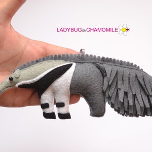 Puede incluir: Adorno de oso hormiguero hecho a mano en fieltro en tonos grises, blancos y negros. El oso hormiguero tiene un hocico largo, ojos negros peque&ntilde;os y una cola con flecos. El texto "LADYBUGONCHAMOMILE" es visible en la esquina superior derecha.