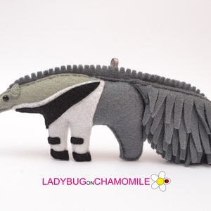 Puede incluir: Adorno de oso hormiguero hecho a mano en fieltro. El oso hormiguero es principalmente gris, con una franja blanca y negra en su costado. Tiene un hocico largo, ojos negros peque&ntilde;os y una cola con flecos. El texto "LADYBUGonCHAMOMILE" est&aacute; en la parte inferior.