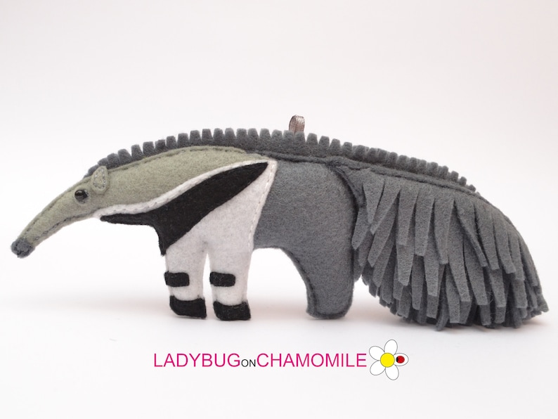 Puede incluir: Adorno de oso hormiguero hecho a mano en fieltro. El oso hormiguero es principalmente gris, con una franja blanca y negra en su costado. Tiene un hocico largo, ojos negros peque&ntilde;os y una cola con flecos. El texto "LADYBUGonCHAMOMILE" est&aacute; en la parte inferior.