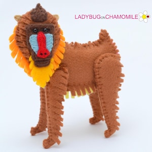 May include: A brown felt stuffed animal resembling a mandrill monkey with a yellow and orange mane, blue nose, and red lips. The animal has stitched seams and is standing on a white surface.
