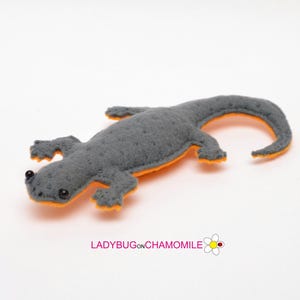 May include: A gray felt lizard toy with orange accents. The lizard has a detailed, textured body, small black eyes, and is lying on a white surface. The underside and edges are bright orange. The text "LADYBUGonCHAMOMILE" is visible.
