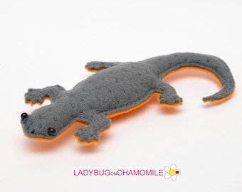Può includere: Un giocattolo a forma di lucertola in feltro grigio con dettagli arancioni. La lucertola ha un corpo strutturato, piccoli occhi neri ed è posizionata su una superficie bianca. Il lato inferiore e i bordi sono arancioni. Il testo "LADYBUGonCHAMOMILE" è visibile.
