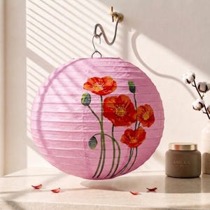 May include: A spherical pink paper lantern featuring a hand-painted design of vibrant red poppy flowers with green stems and buds. The lantern is suspended from a metal hook and displays a ribbed texture characteristic of traditional paper construction.