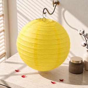 May include: A bright yellow spherical paper lantern with horizontal ribbed detailing, suspended from a curved metal wall-mounted hook. The lantern features a thin wire frame structure visible through the paper material.
