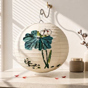 May include: A round paper lantern featuring a hand-painted ink illustration of a teal lotus leaf and a white lotus flower. The lantern has a ribbed texture and is suspended from a silver-toned metal hook. The design includes green stems.