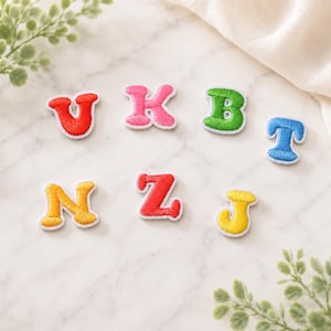 May include: A collection of seven individual embroidered alphabet letter patches in various colors including red, pink, green, blue, and yellow. Each letter features a white stitched border and a textured thread finish, arranged in two rows.