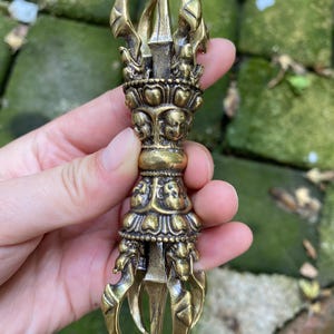 Puede incluir: Un objeto ritual vajra de latón con un mango central que presenta rostros tallados y puntas simétricas que se extienden desde ambos extremos. El metal tiene un acabado dorado pulido con detalles intrincados en los rostros y puntas.