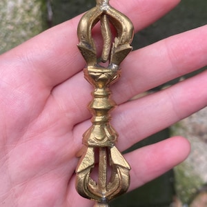 Puede incluir: Un objeto ritual vajra de metal dorado con un diseño simétrico, un mango central y extremos con puntas. El artículo tiene un acabado pulido con detalles intrincados en las puntas y una terminación en punta en ambos extremos superior e inferior.