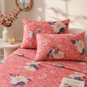 Puede incluir: Un juego de cama color coral que incluye dos almohadas rectangulares y una sábana a juego. La tela está decorada con un patrón repetitivo de margaritas blancas, pájaros azules y rojos y follaje blanco delicado en toda la superficie.