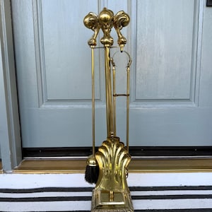 May include: A polished brass fireplace tool set featuring a central stand with a claw-foot base, a shovel with a shell-shaped design, a poker, and a brush with black bristles. The set includes a vertical rack to hold the tools securely in place.
